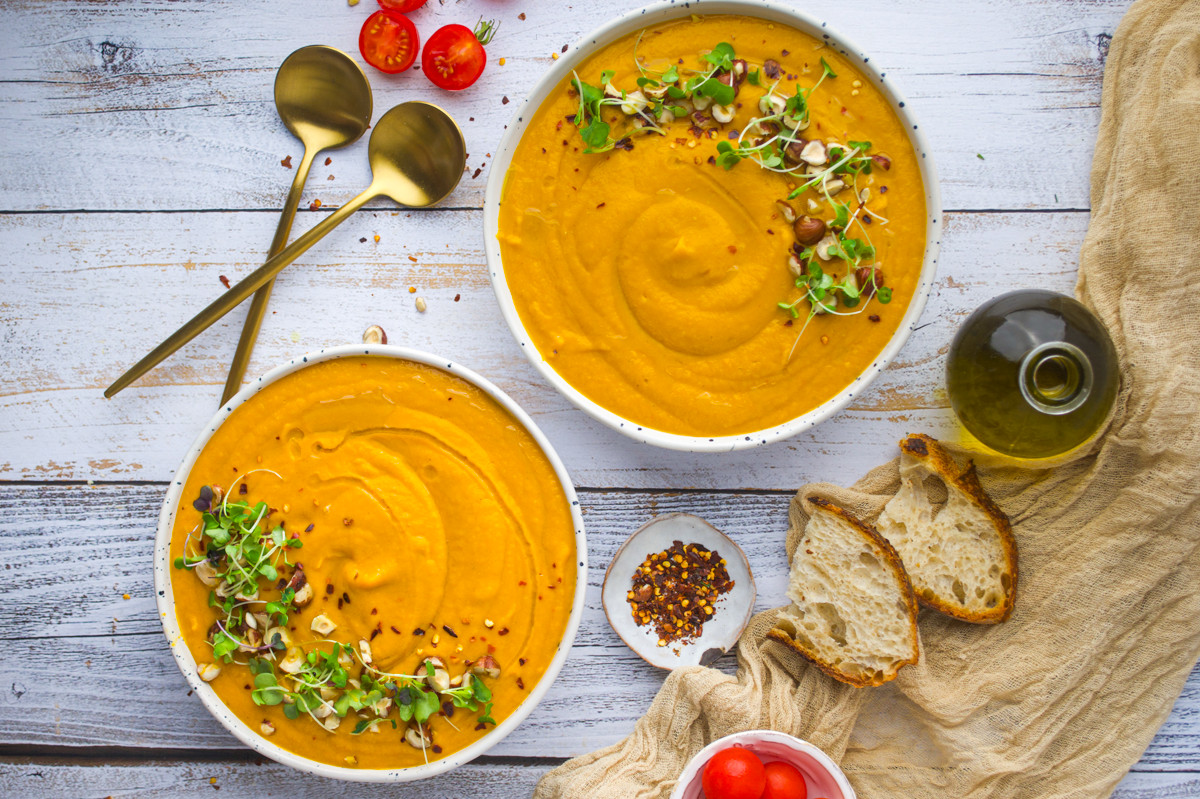Two bowls of creamy orange soup, garnished with microgreens and seeds, sit on a rustic wooden table with bread, olive oil, cherry tomatoes, gold spoons, and a small dish of chili flakes nearby.