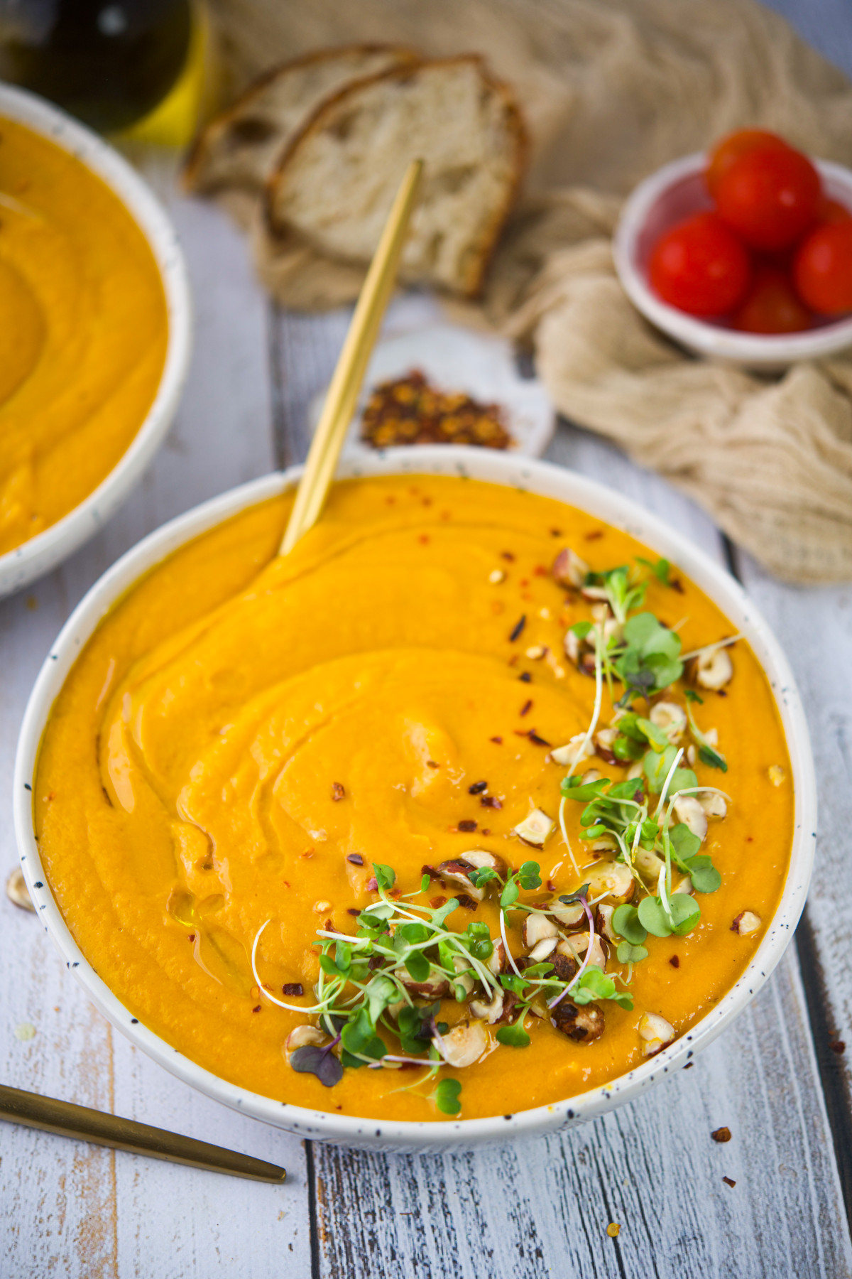 A bowl of creamy orange soup topped with microgreens and chopped nuts, with bread and cherry tomatoes in the background on a rustic white table.