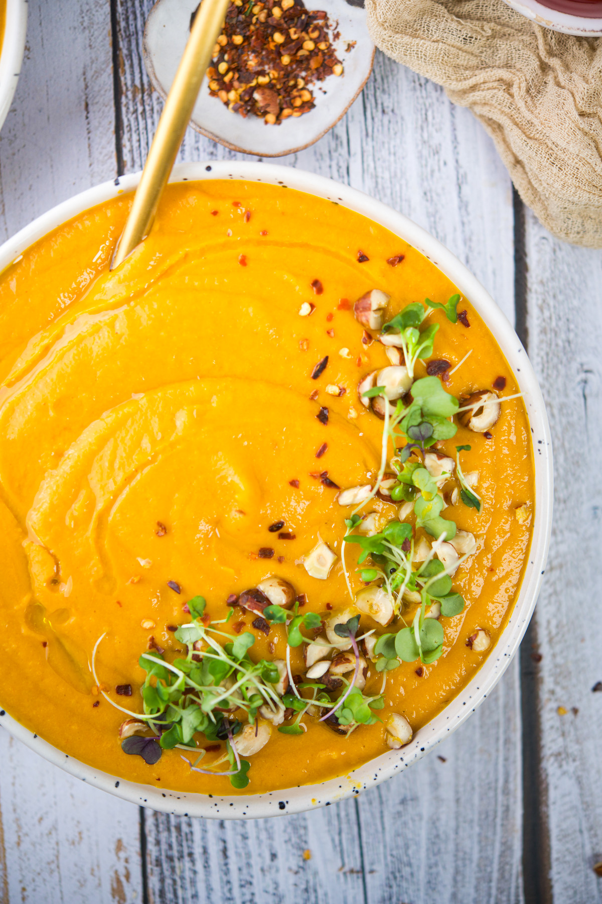 A bowl of creamy orange soup topped with chopped nuts, microgreens, and red pepper flakes, with a spoon resting inside. The bowl sits on a rustic white wooden surface.