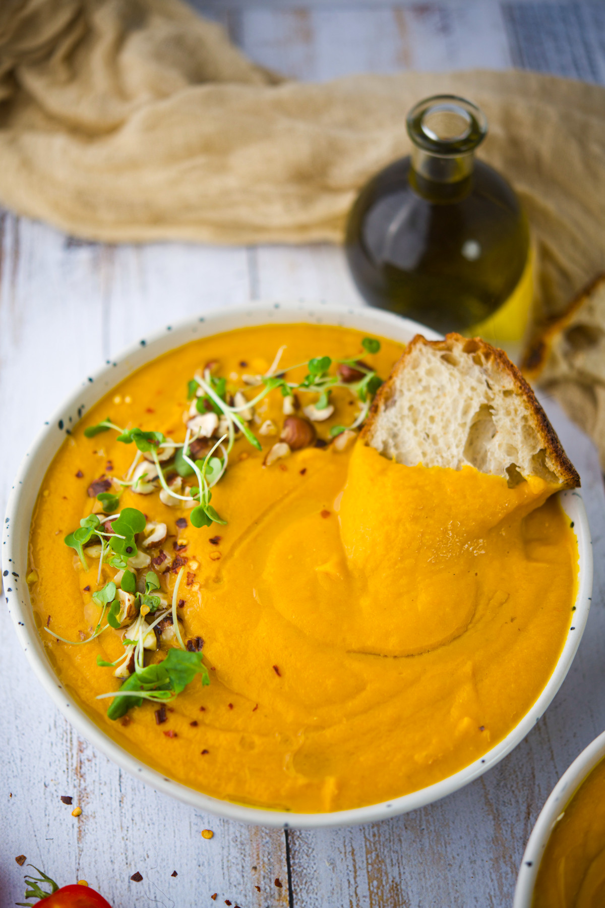 Bowl of creamy orange soup garnished with microgreens and chopped nuts, with a slice of crusty bread dipped in. A bottle of olive oil and beige cloth are in the background on a white wooden table.