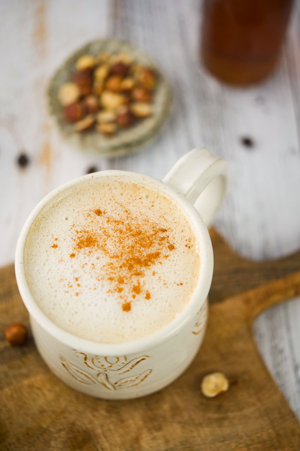 A creamy hazelnut latte topped with cinnamon in a white mug sits on a wooden board, with whole hazelnuts scattered nearby and more hazelnuts in a small dish in the background.