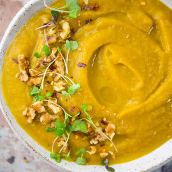A bowl of roasted sweet potato pear soup topped with microgreens, chopped walnuts, and red pepper flakes.