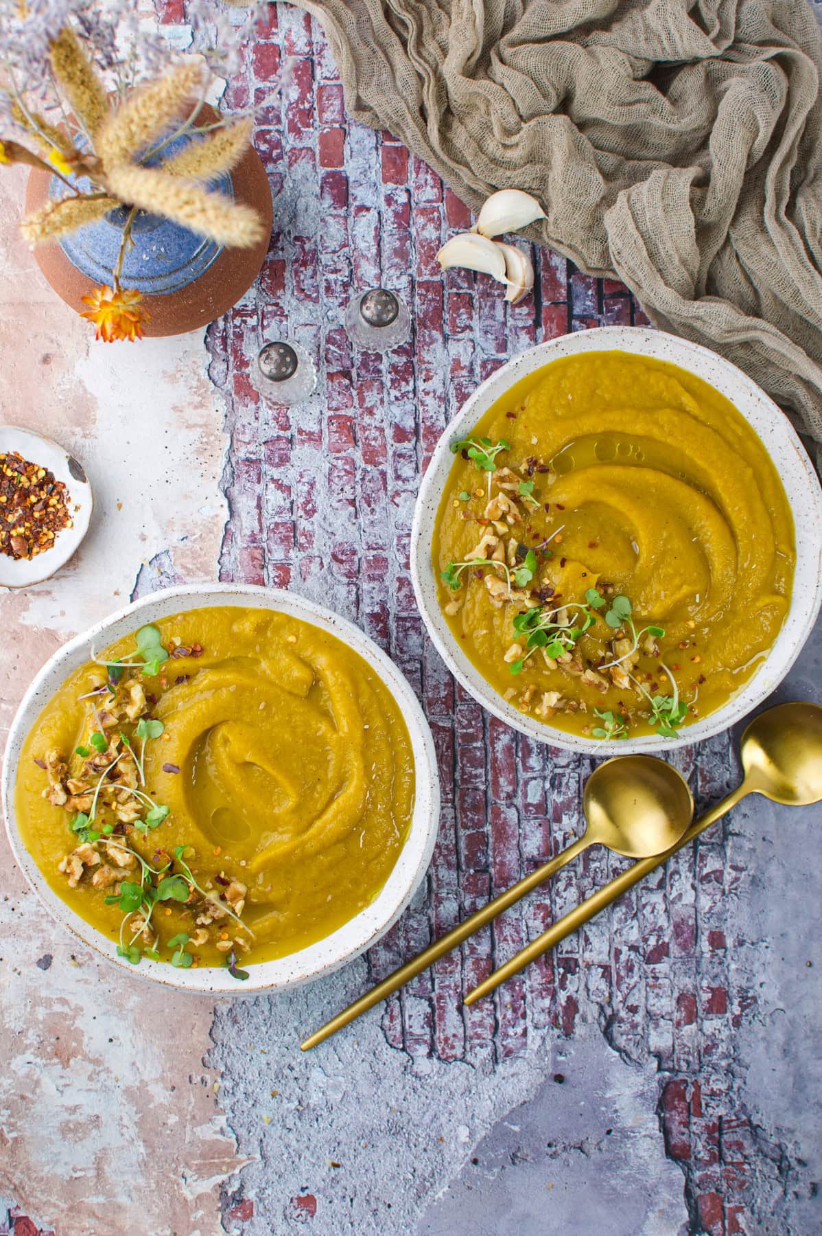 Two bowls of thick yellow soup topped with microgreens and nuts sit on a rustic textured surface, beside two gold spoons, a dish of chili flakes, garlic cloves, and a beige cloth.