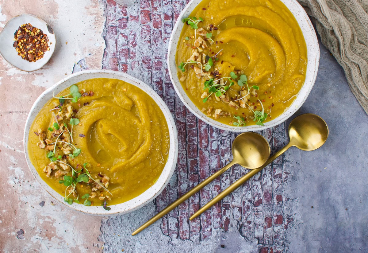 Two bowls of creamy soup, garnished with microgreens and walnuts, sit on a textured surface beside two gold spoons. A small dish of red pepper flakes is nearby. The scene is cozy and inviting.