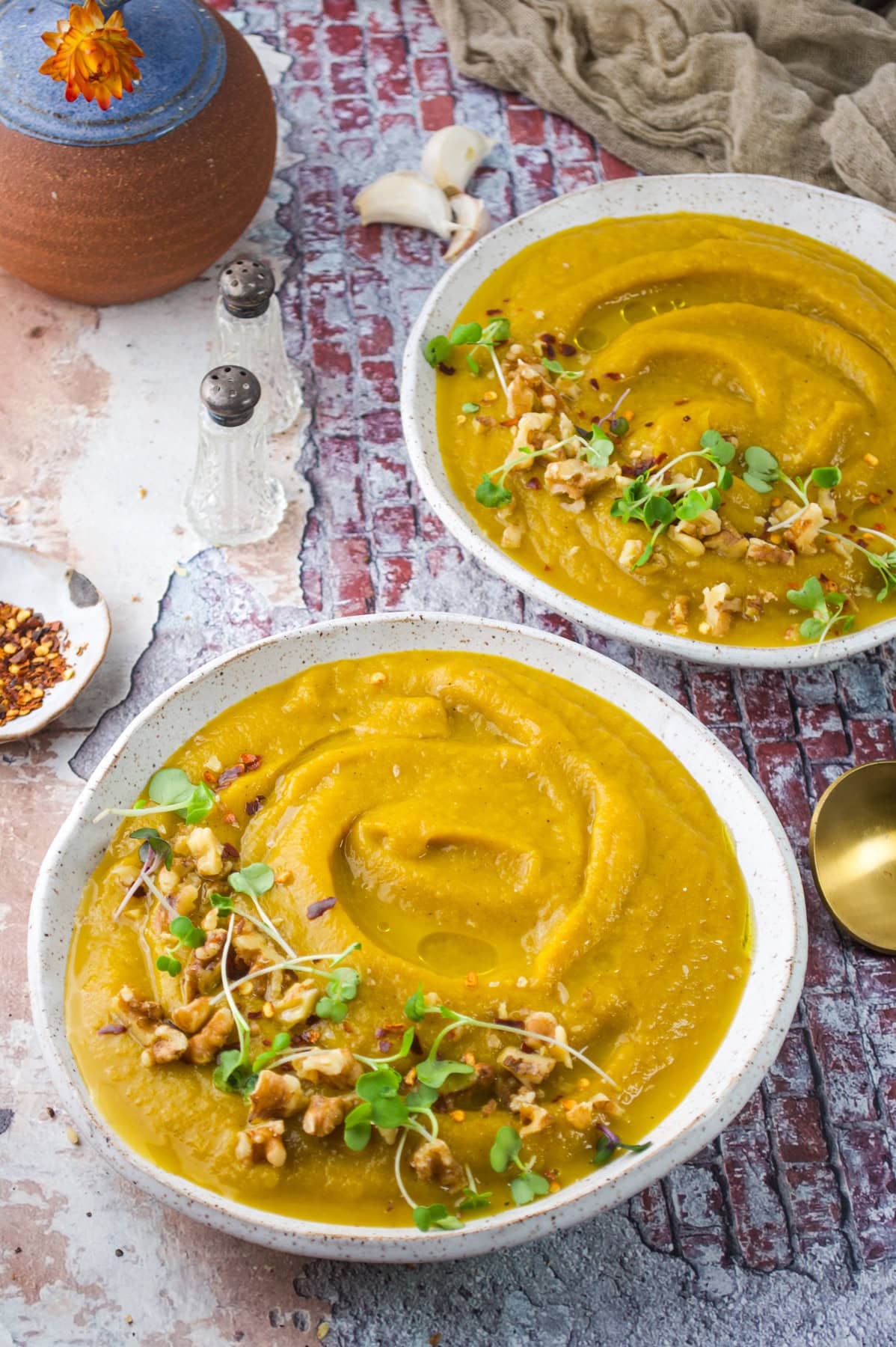 Two bowls of creamy yellow soup topped with microgreens, walnuts, and red pepper flakes sit on a rustic table beside a gold spoon, salt and pepper shakers, and a small clay pot with an orange flower.