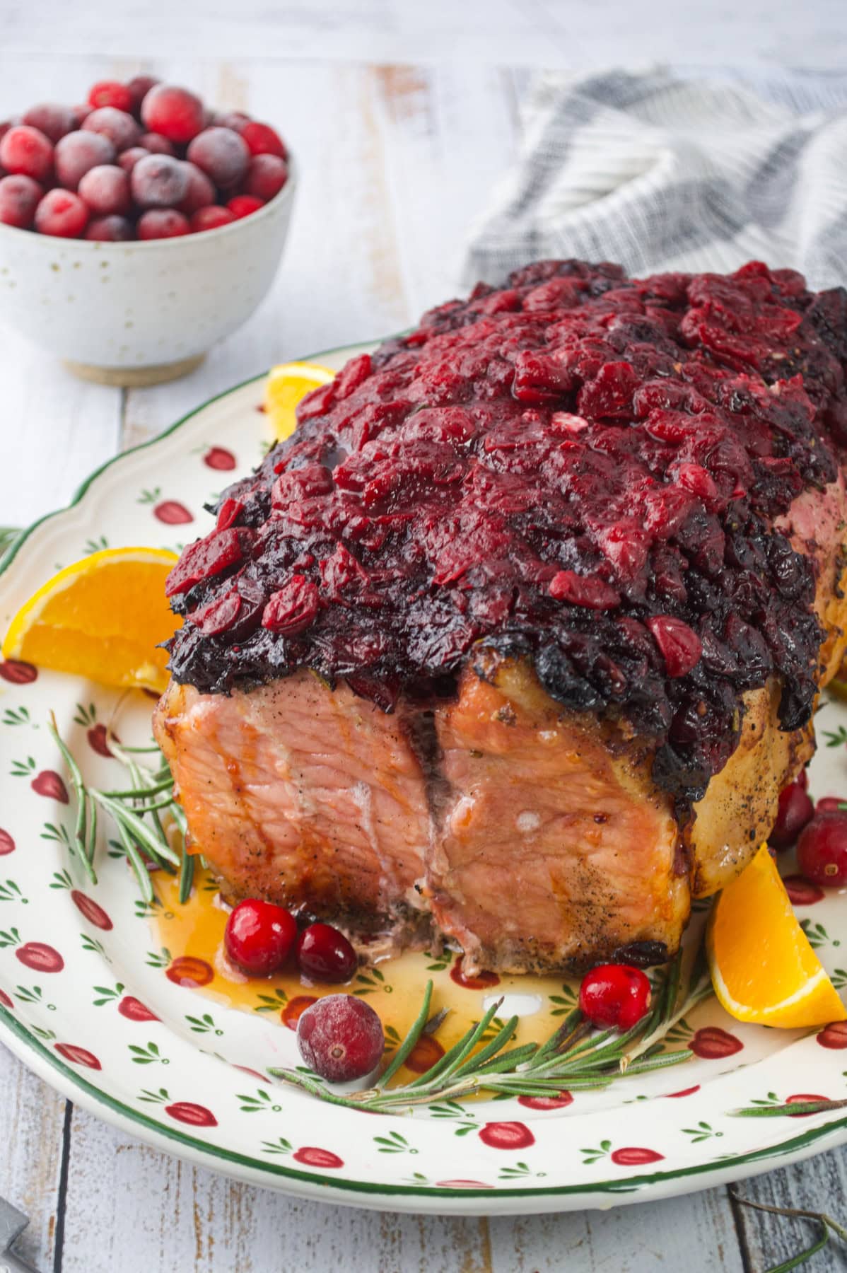 A roasted ham topped with a chunky cranberry glaze sits on a decorative platter, garnished with fresh rosemary, orange slices, and cranberries. A bowl of cranberries and a napkin are in the background.