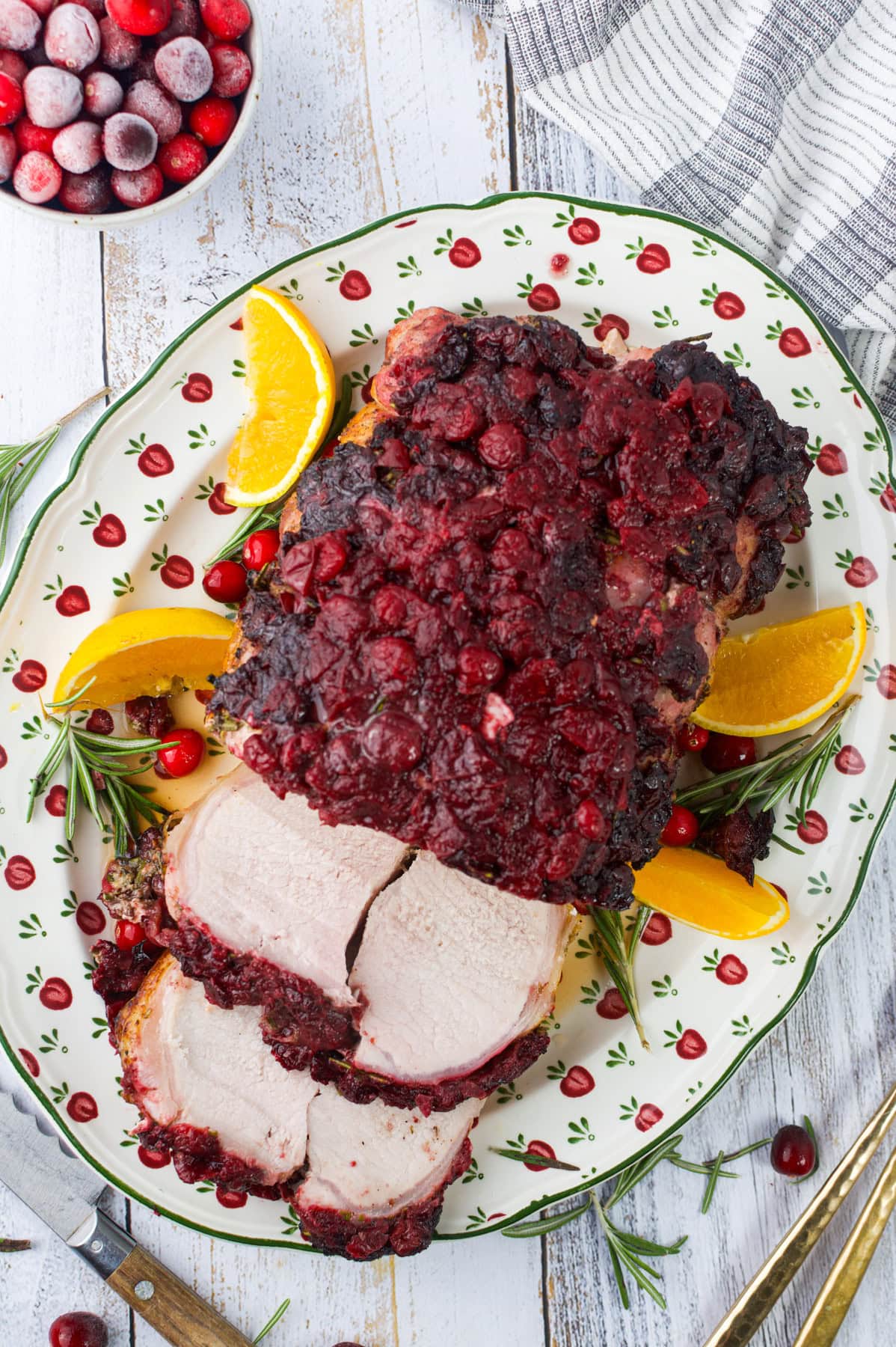 A platter with a cranberry-glazed pork roast, partially sliced, garnished with orange wedges, fresh cranberries, and rosemary sprigs. A bowl of cranberries and a striped towel are nearby on a white wooden table.