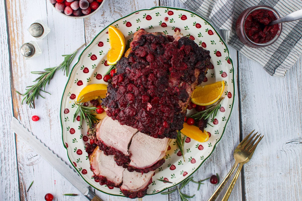 A platter of roast pork topped with cranberry sauce, garnished with orange slices, rosemary, and cranberries, sits on a white wooden table with a knife, fork, napkin, and a small jar of cranberry sauce nearby.