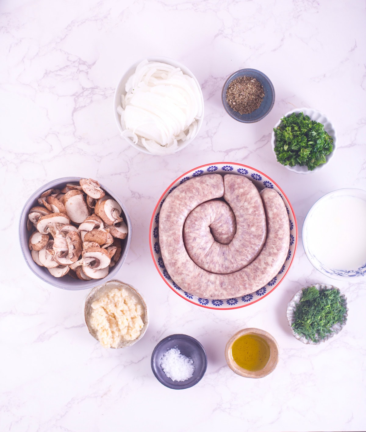 A flat lay of raw sausage in a bowl surrounded by small bowls of sliced onions, mushrooms, grated cheese, chopped herbs, ground pepper, salt, olive oil, and a bowl of cream on a white marble surface.