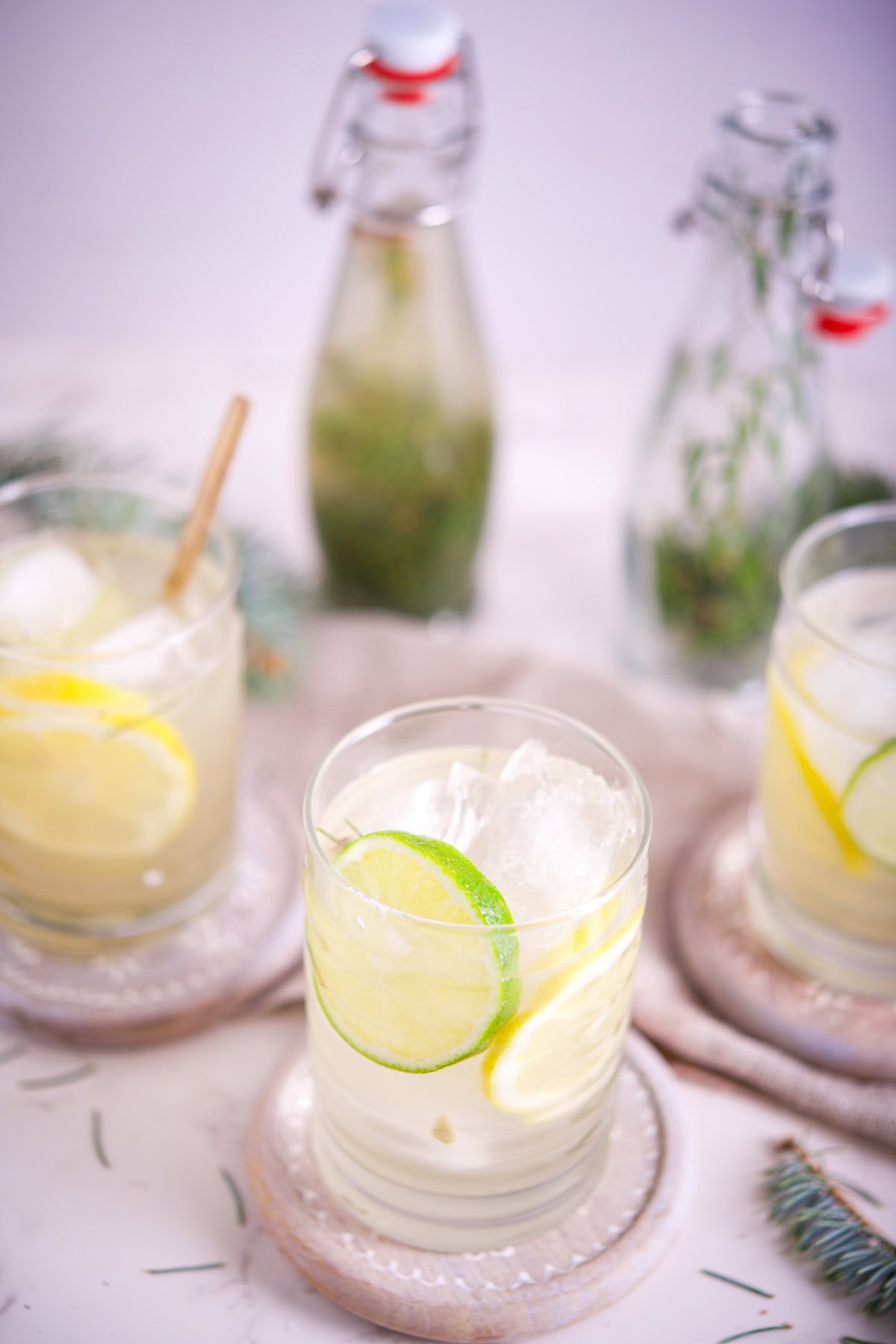 Three glasses of iced lemon and lime water sit on coasters, garnished with citrus slices. Behind them are two glass bottles with herbs inside, all arranged on a light-colored surface with scattered pine needles.