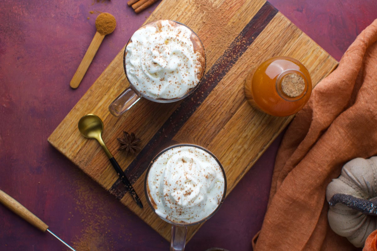 Top view of two mugs of whipped cream-topped drinks on a wooden board, with a bottle of syrup, a gold spoon, cinnamon sticks, and a rust-colored cloth on a reddish surface.