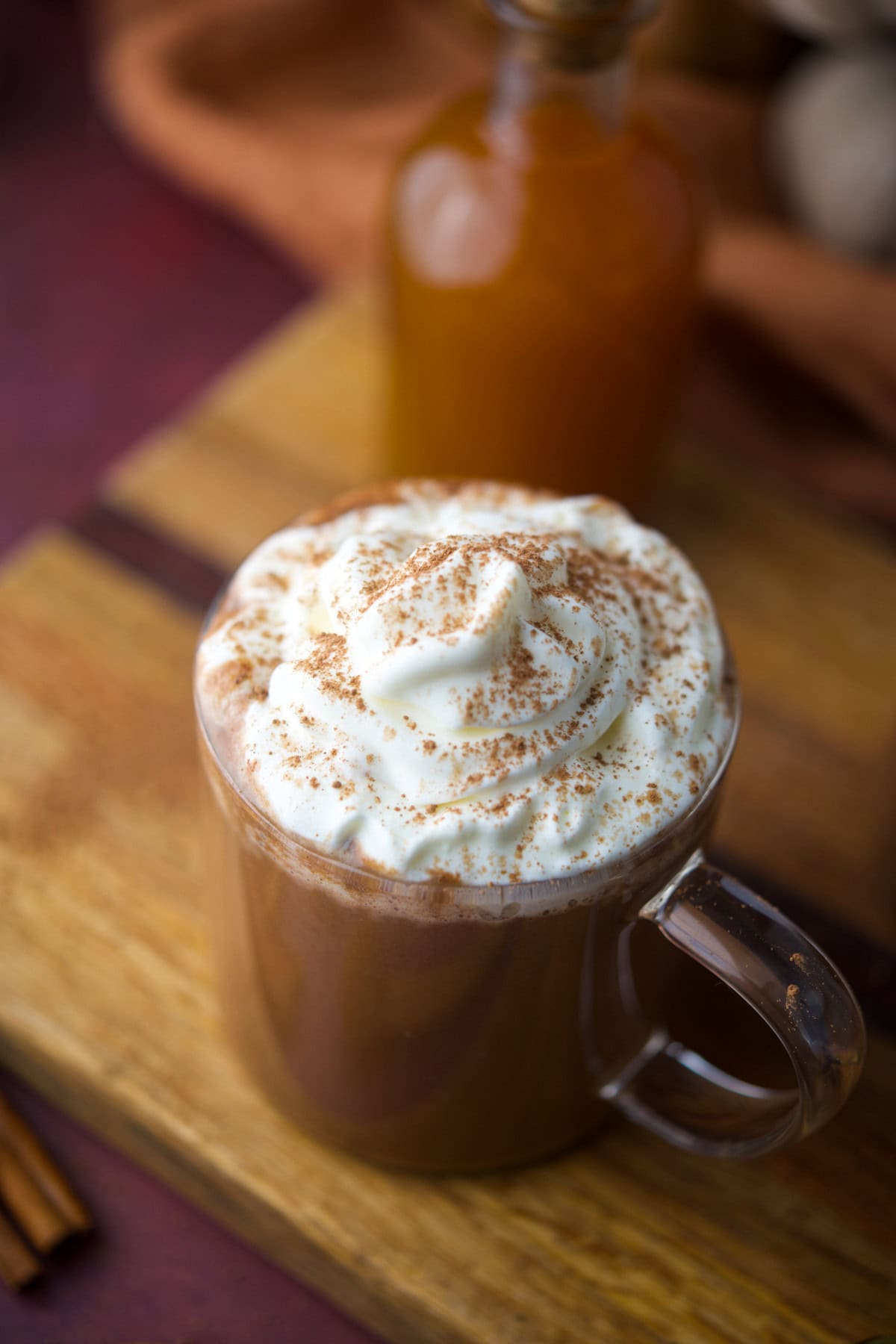 A clear glass mug filled with Chocolate Pumpkin Spiced Latte, topped with whipped cream and a sprinkle of cinnamon, placed on a wooden surface with a glass bottle in the background.