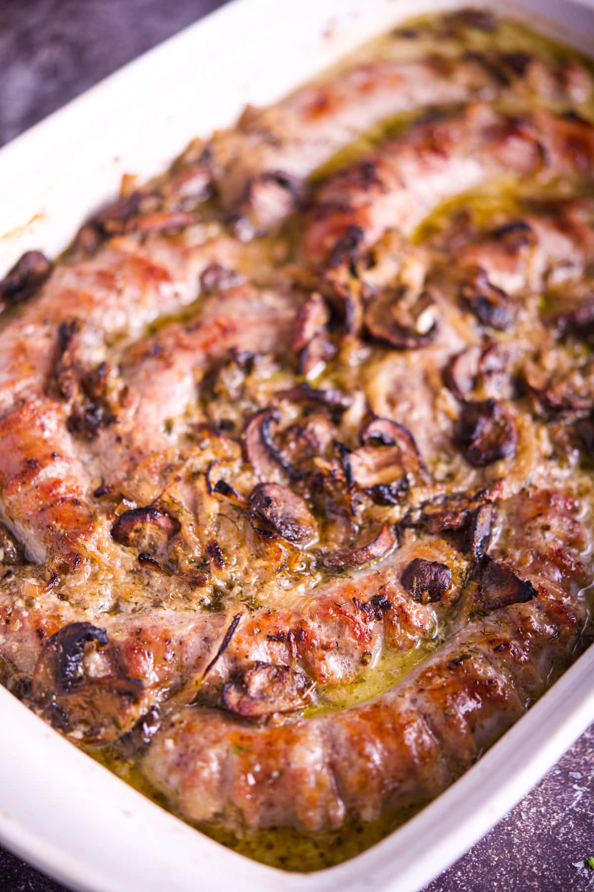 Close-up of browned sausages baked in a white dish, topped with sliced mushrooms and herbs, sitting in a golden, bubbly sauce.