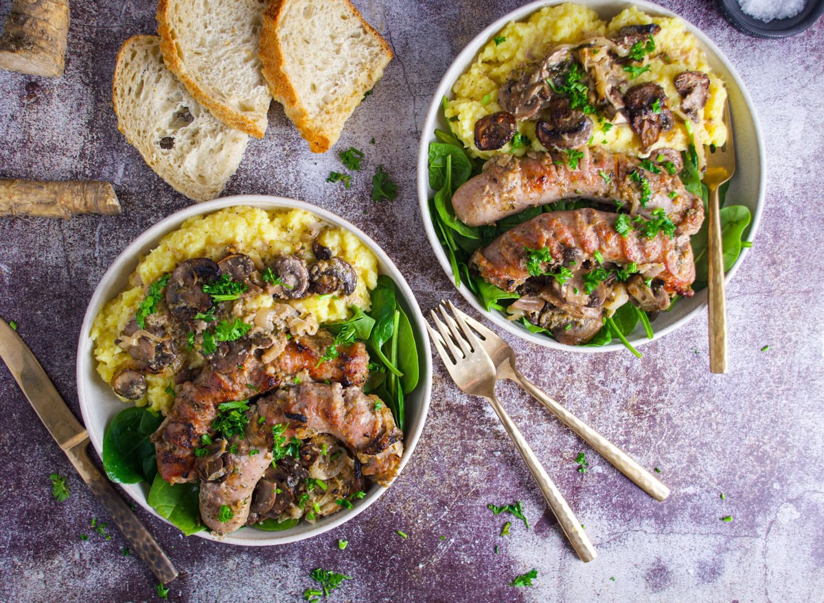 Two bowls of creamy mashed potatoes topped with grilled sausages, sautéed mushrooms, and fresh parsley, with spinach on the side. Slices of rustic bread and two forks are placed nearby on a textured surface.