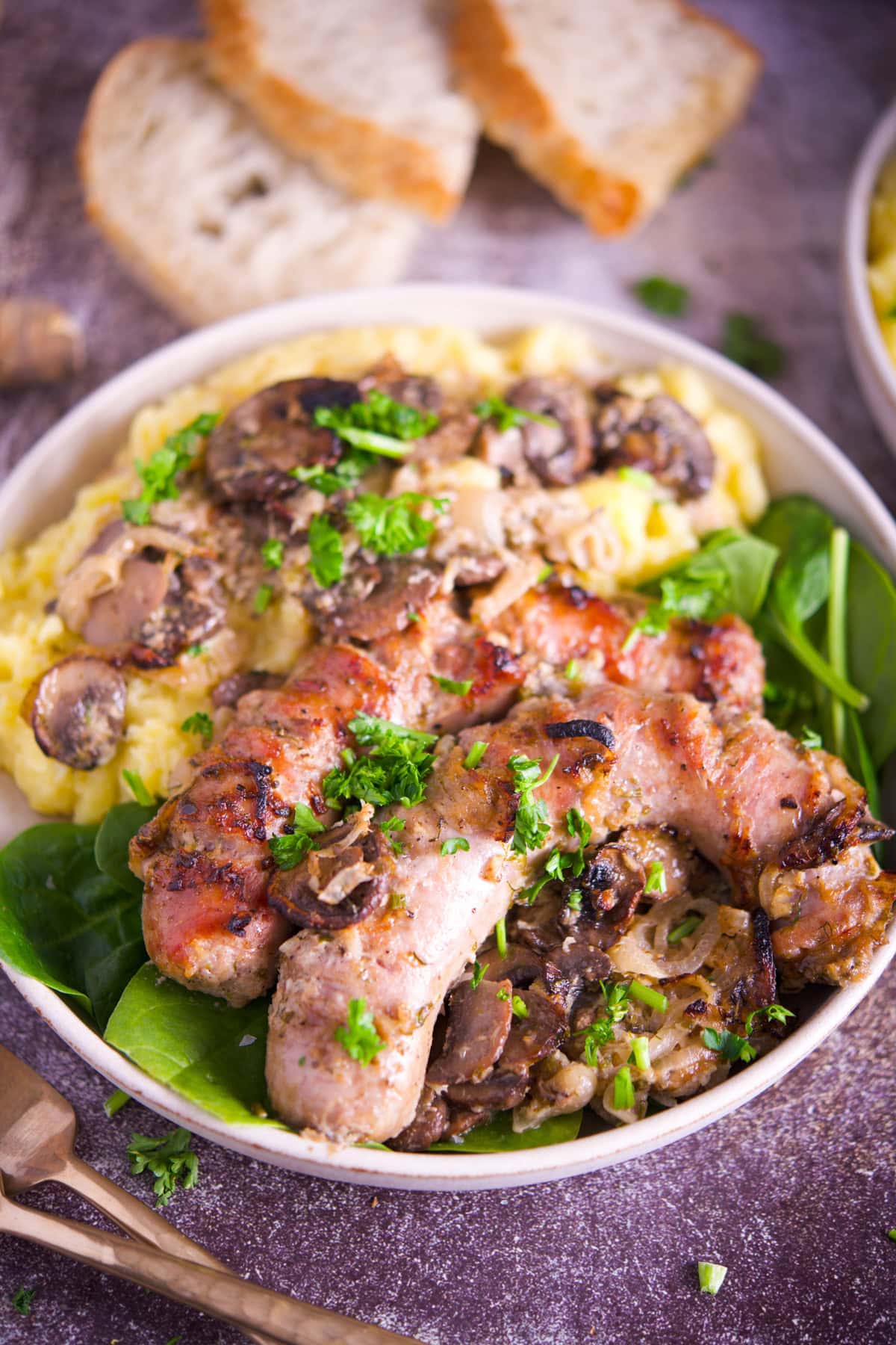 A bowl of grilled sausages with sautéed mushrooms and fresh parsley, served over creamy mashed potatoes and spinach. Slices of bread are in the background.
