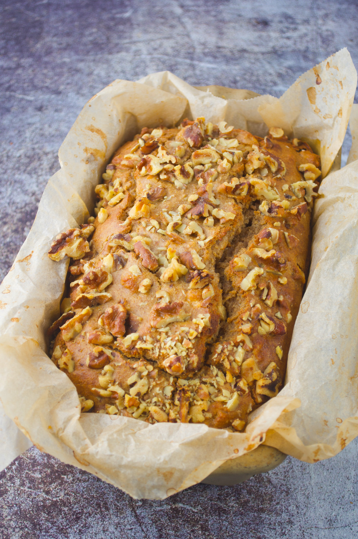A loaf of banana bread topped with chopped walnuts sits in a parchment-lined baking pan on a textured surface.