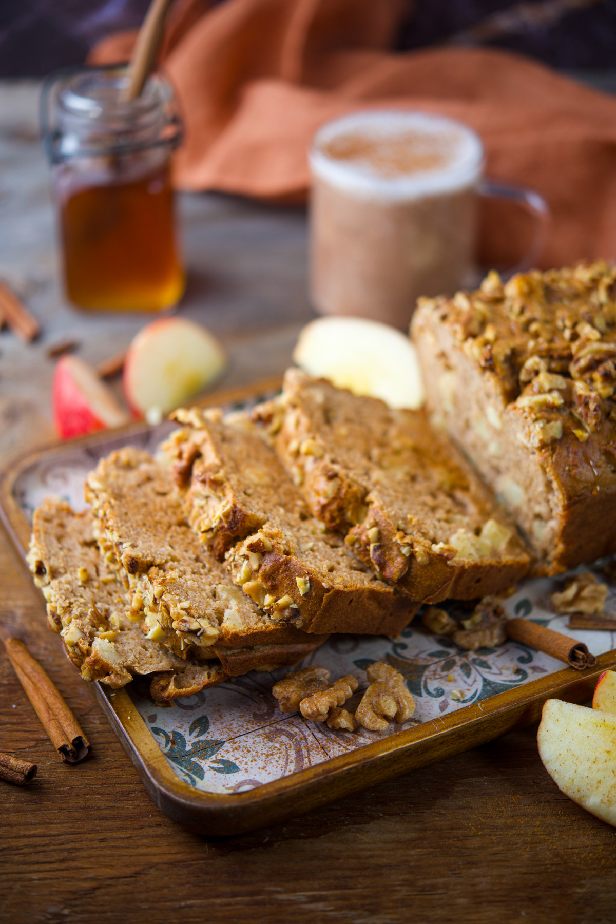 Sliced apple bread with walnuts on a decorative tray, surrounded by apple slices and cinnamon sticks. In the background, theres a glass of honey and a mug with a foamy drink.