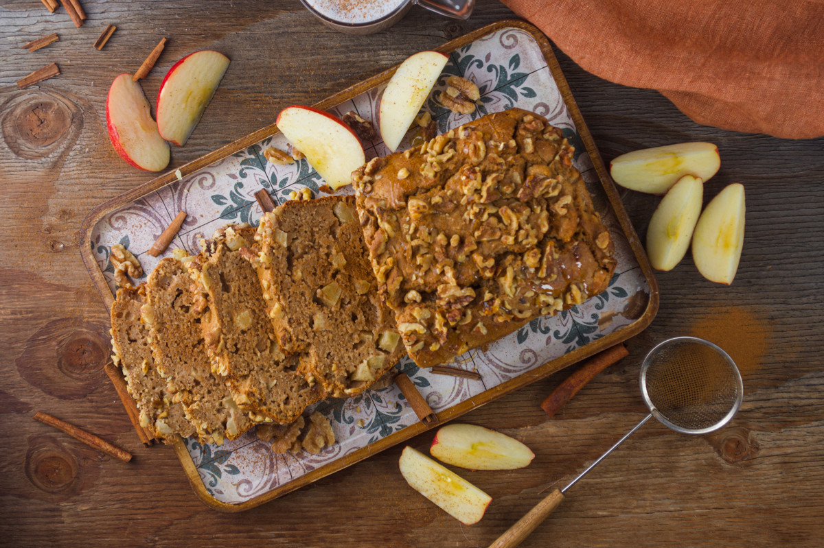 A loaf of apple walnut bread, partially sliced, is on a decorative tray with apple slices and walnuts. Cinnamon sticks and a sifter rest nearby on a wooden table.