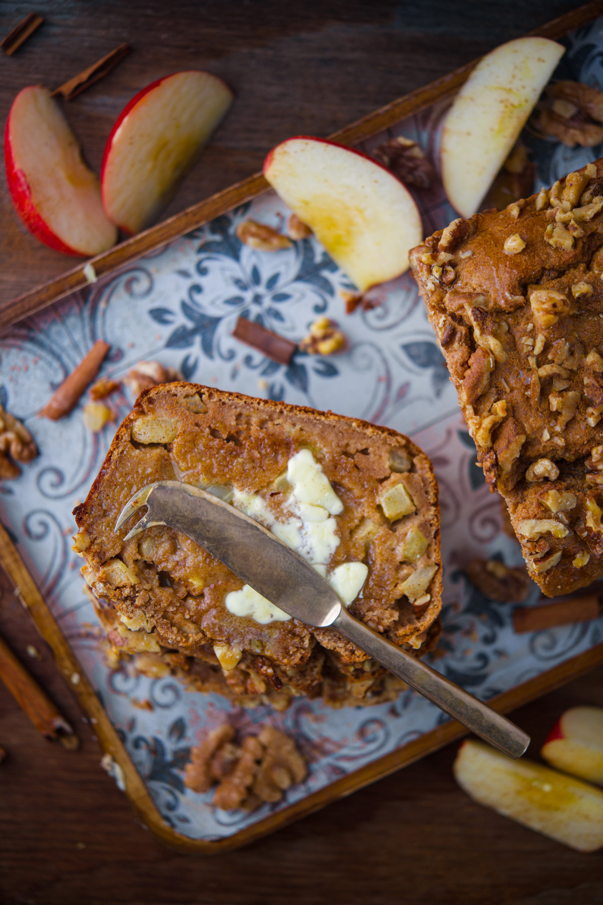 A sliced loaf of apple bread with walnuts on top, served with butter and a butter knife. Apple slices and walnuts are scattered around on a decorative tray.