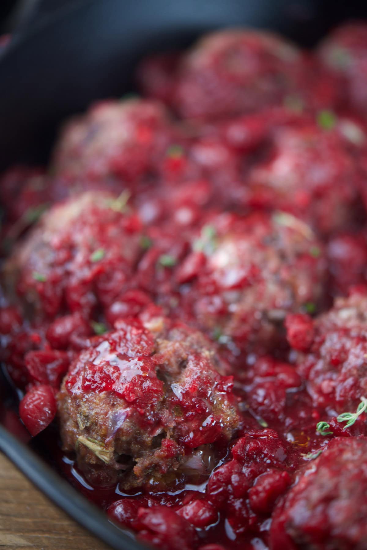 Meatballs topped with a chunky red raspberry sauce, garnished with small green herb leaves, served in a dark dish.