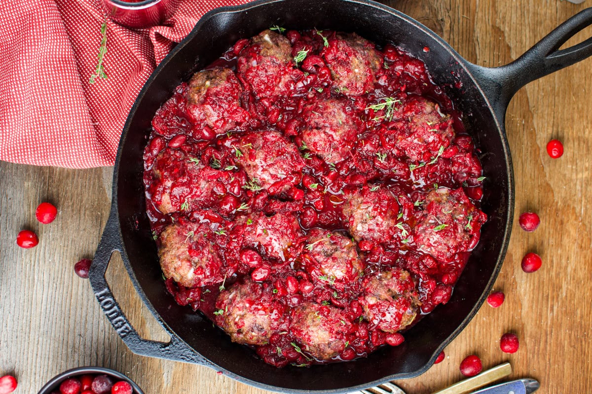 A cast iron skillet filled with meatballs covered in a chunky red cranberry sauce, garnished with herbs. The skillet sits on a wooden surface with scattered cranberries and a red cloth nearby.