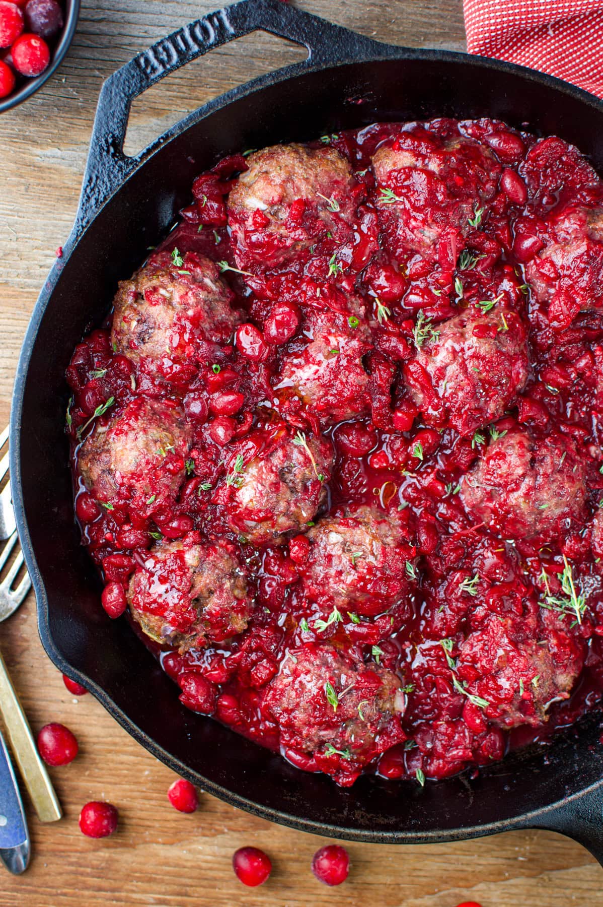 A cast iron skillet filled with meatballs covered in a chunky, vibrant red cranberry sauce, garnished with fresh herbs, sits on a wooden table next to cutlery and a red napkin.