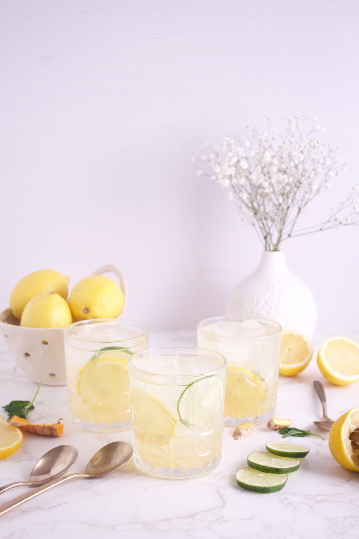 Three glasses of lemon-lime water with ice and slices of lemon and cucumber on a marble surface, surrounded by fresh lemons, a vase with white flowers, spoons, and lemon and cucumber slices.