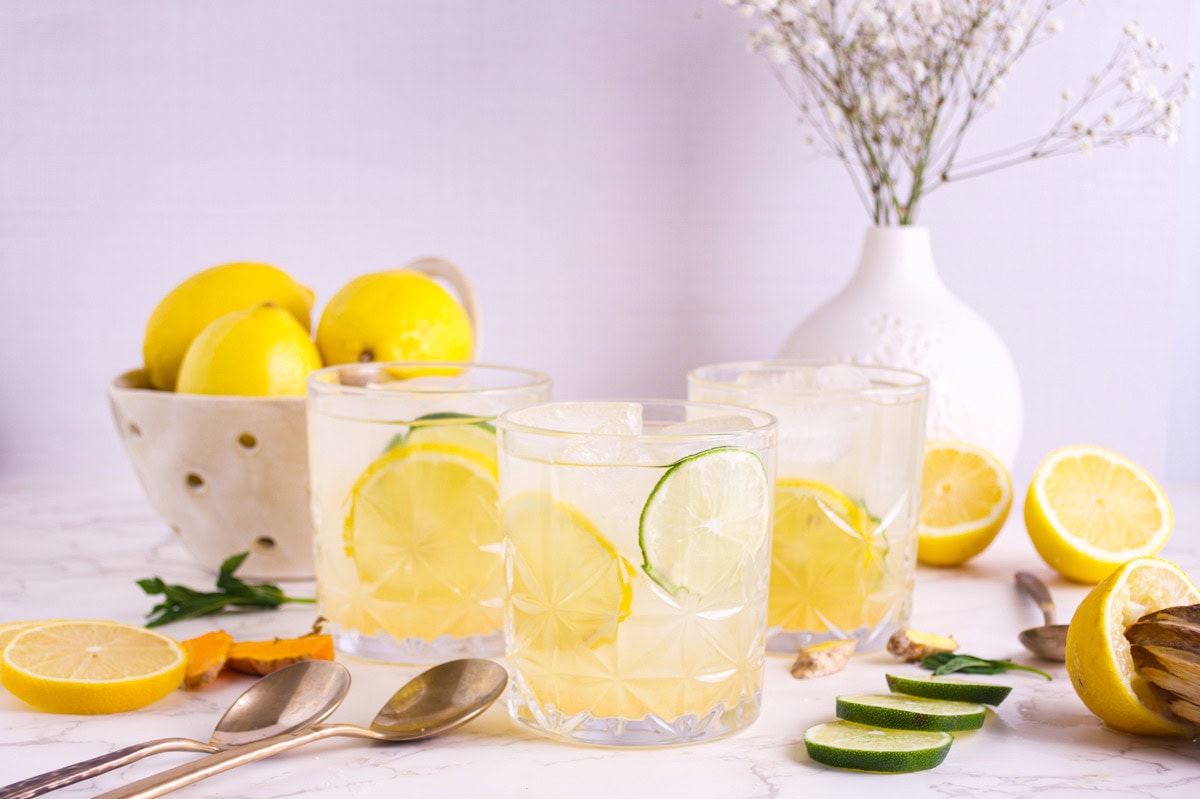 Three glasses of lemonade with lemon and lime slices on a white table, surrounded by sliced lemons, limes, mint leaves, a bowl of whole lemons, spoons, and a white vase with flowers in the background.