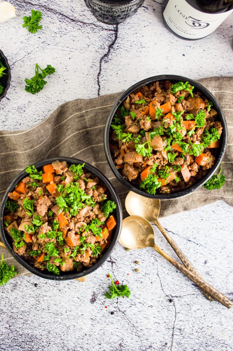 Top-down view of two black bowls filled with ground meat stew featuring carrots, mushrooms, and parsley, placed on a linen cloth with gold spoons, black pepper, and fresh parsley scattered around.