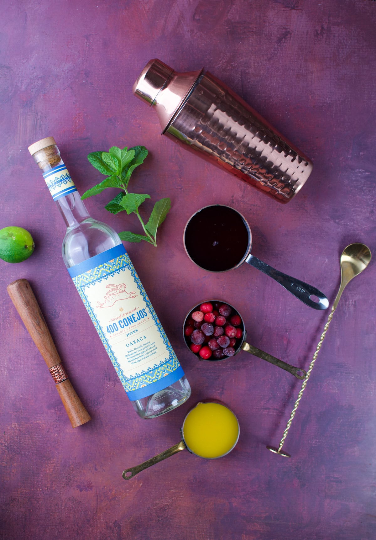 A cocktail shaker, bottle of 400 Conejos mezcal, a jigger, wooden muddler, mint, lime, cup of orange juice, cup of mixed berries, and cup of dark liquid are arranged on a purple surface.