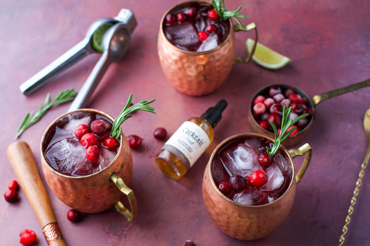 Three copper mugs filled with ice, cranberries, and rosemary sprigs are on a table with cocktail tools, cranberries, a lime wedge, and a small bottle of bitters. The scene has a festive, rustic feel.