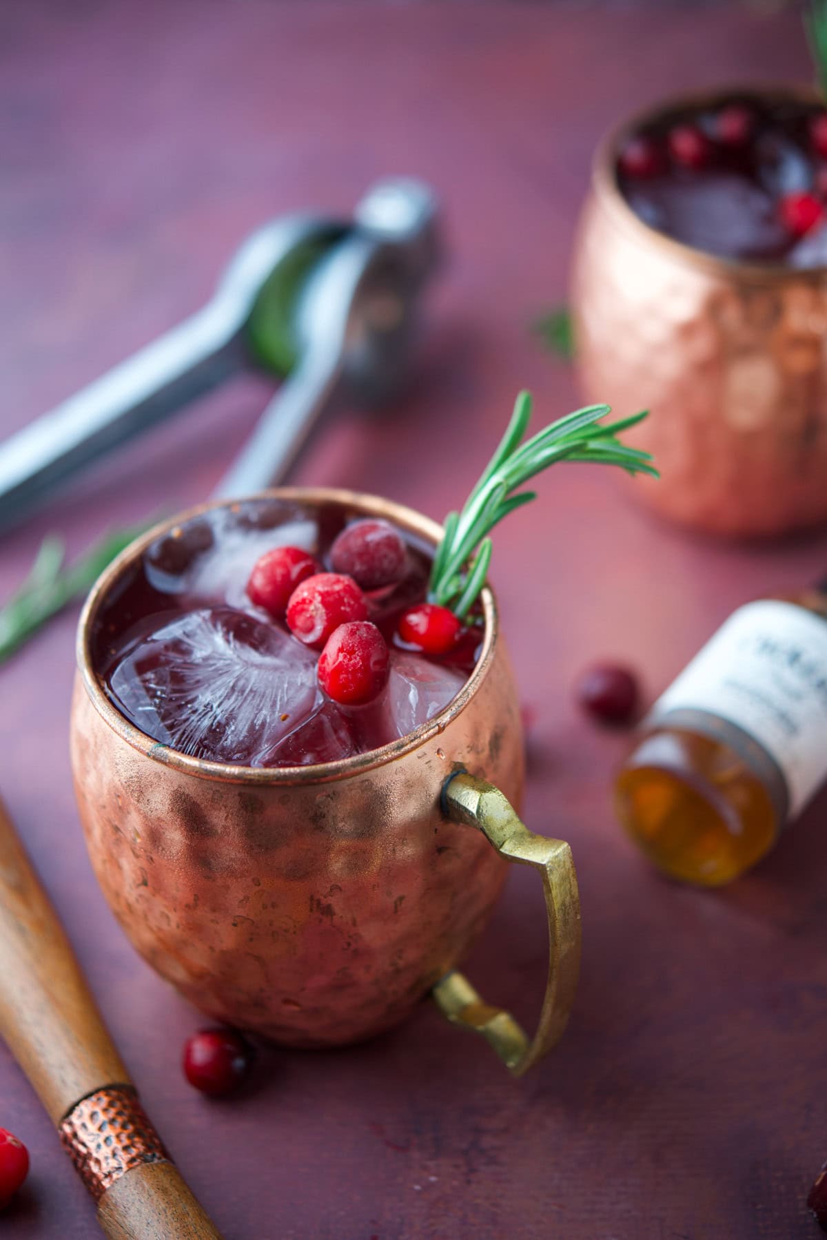 A copper mug filled with ice, cranberries, and a sprig of rosemary sits on a table. Another similar mug, a small bottle, a muddler, and a metal citrus press are nearby.