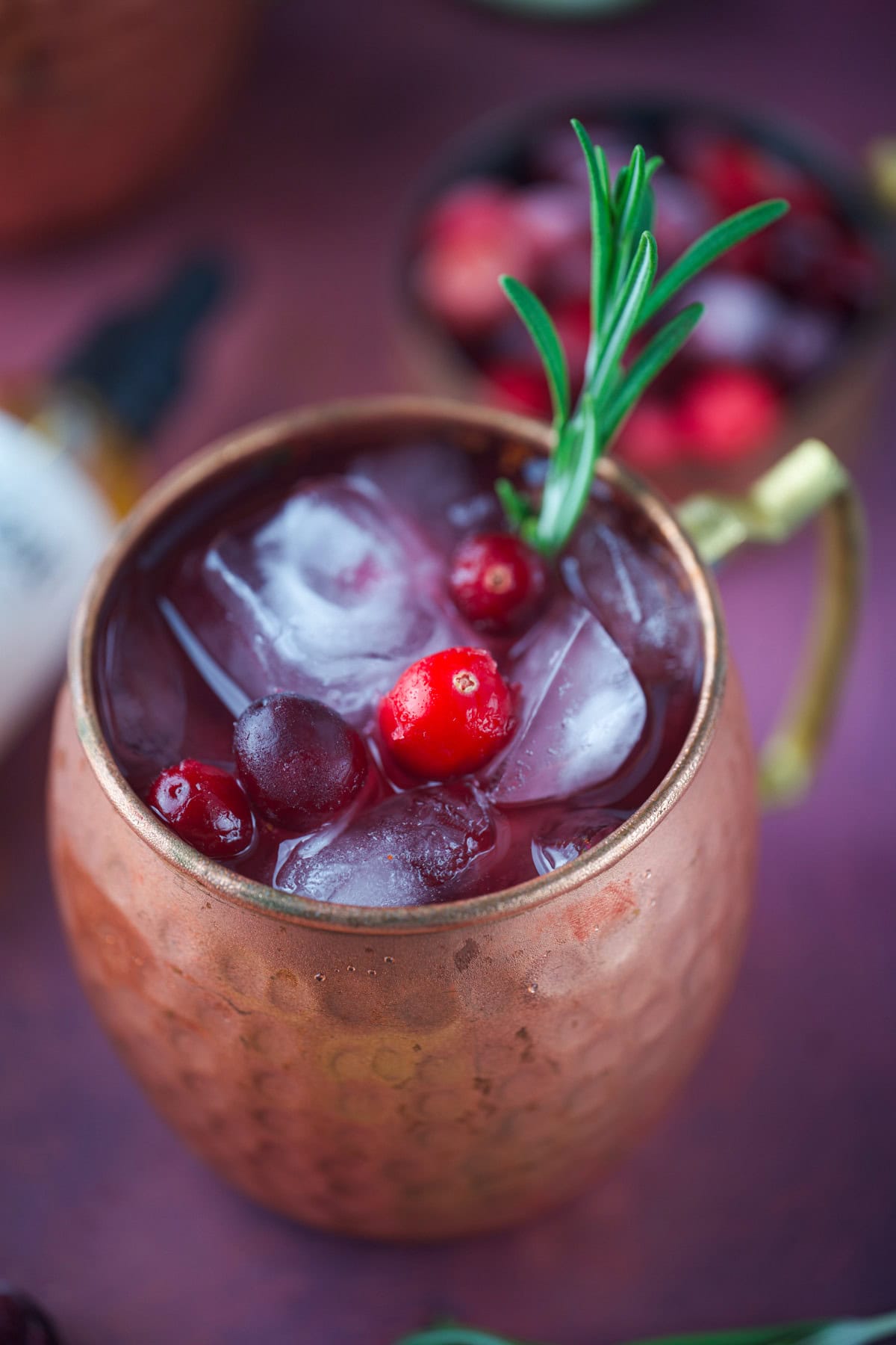 A copper mug filled with a red, iced cocktail, garnished with cranberries and a sprig of rosemary. Blurred cranberries and another drink are visible in the background.