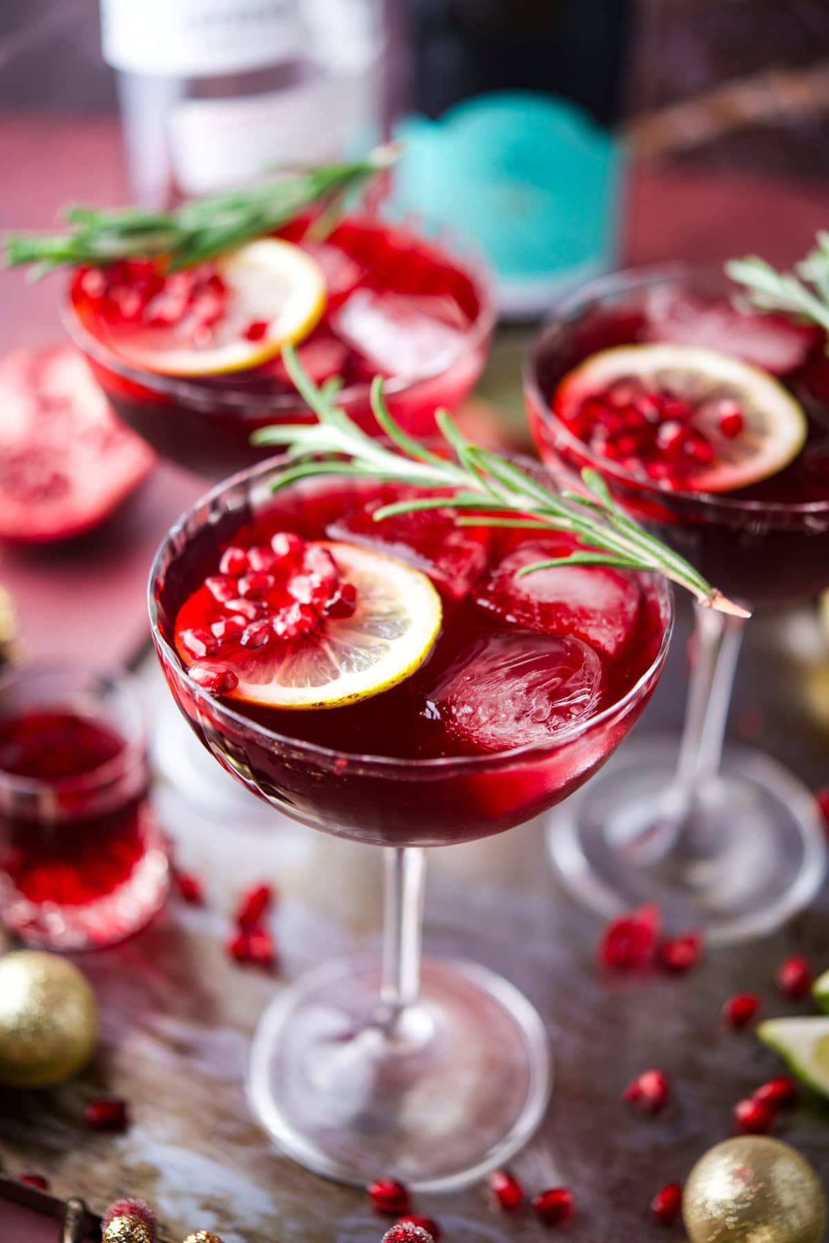 Three glasses of Pomegranate French 75, are garnished with lemon slices, pomegranate seeds, and rosemary sprigs. The drinks are set on a tray with festive decorations and blurred bottles in the background.