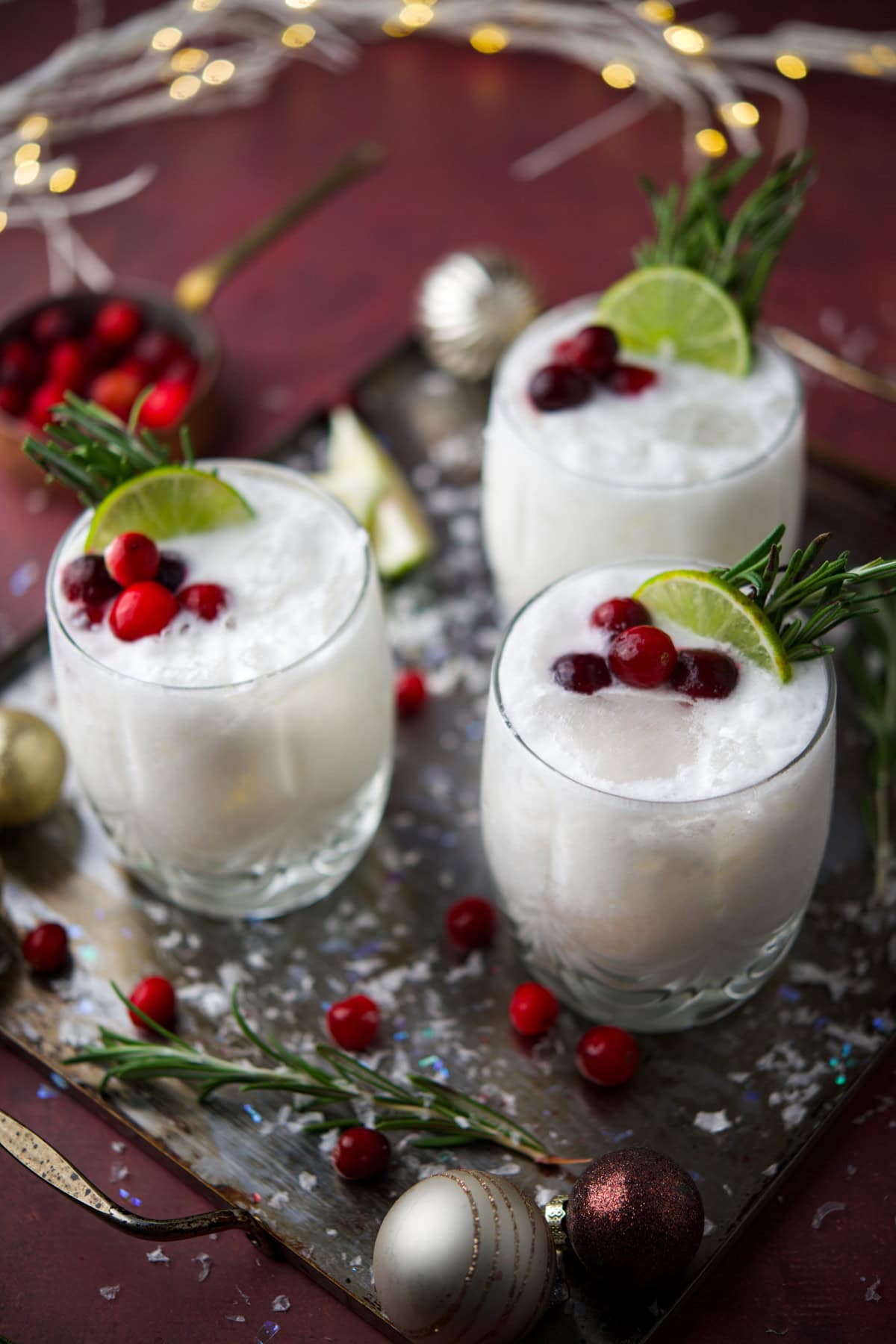 Three frothy white cocktails in glasses are garnished with cranberries, lime slices, and sprigs of rosemary. The drinks sit on a tray with festive decorations, cranberries, rosemary, and Christmas ornaments around them.