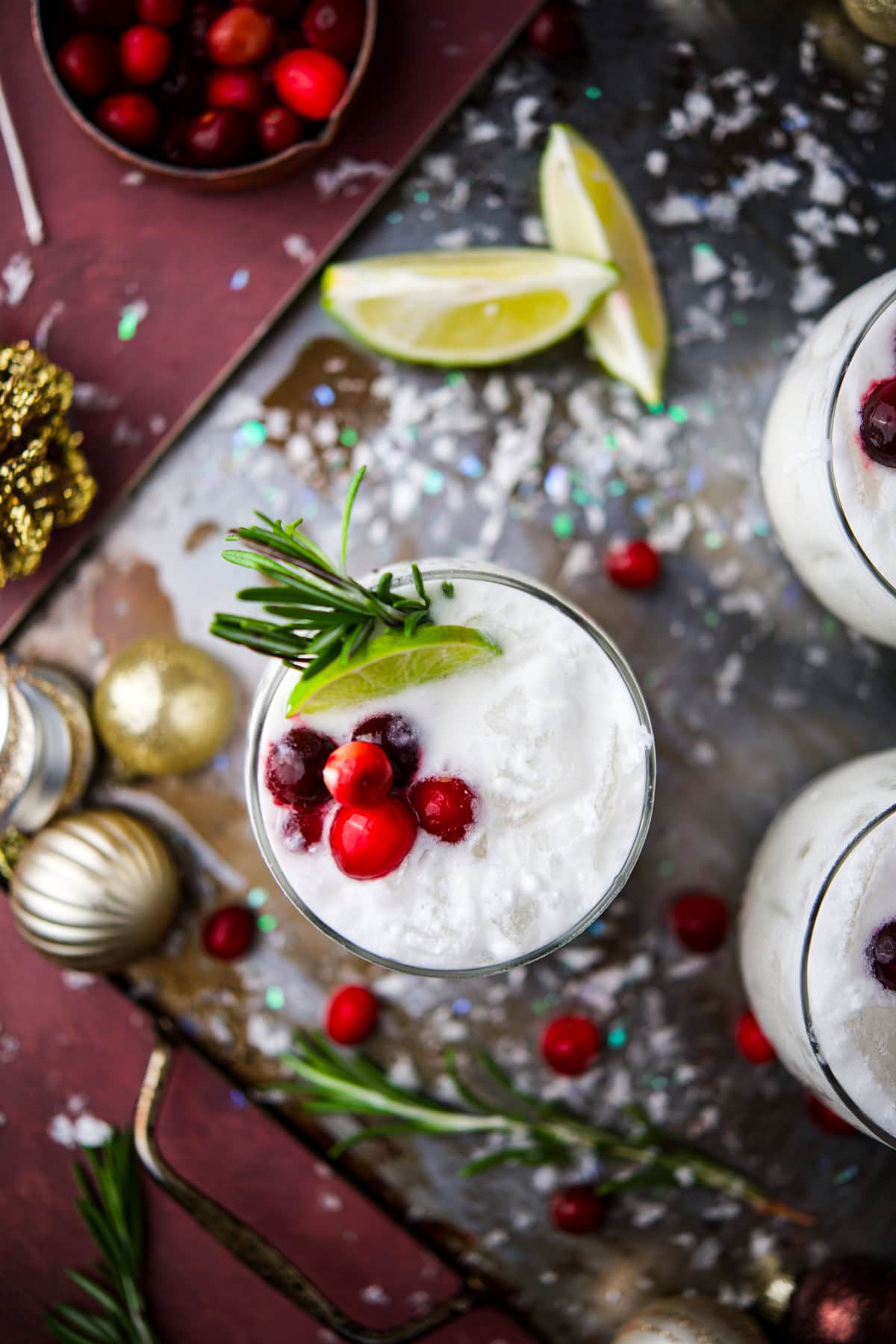 A festive cocktail garnished with cranberries, a sprig of rosemary, and a lime wedge sits on a table with scattered cranberries, lime slices, and holiday decorations.