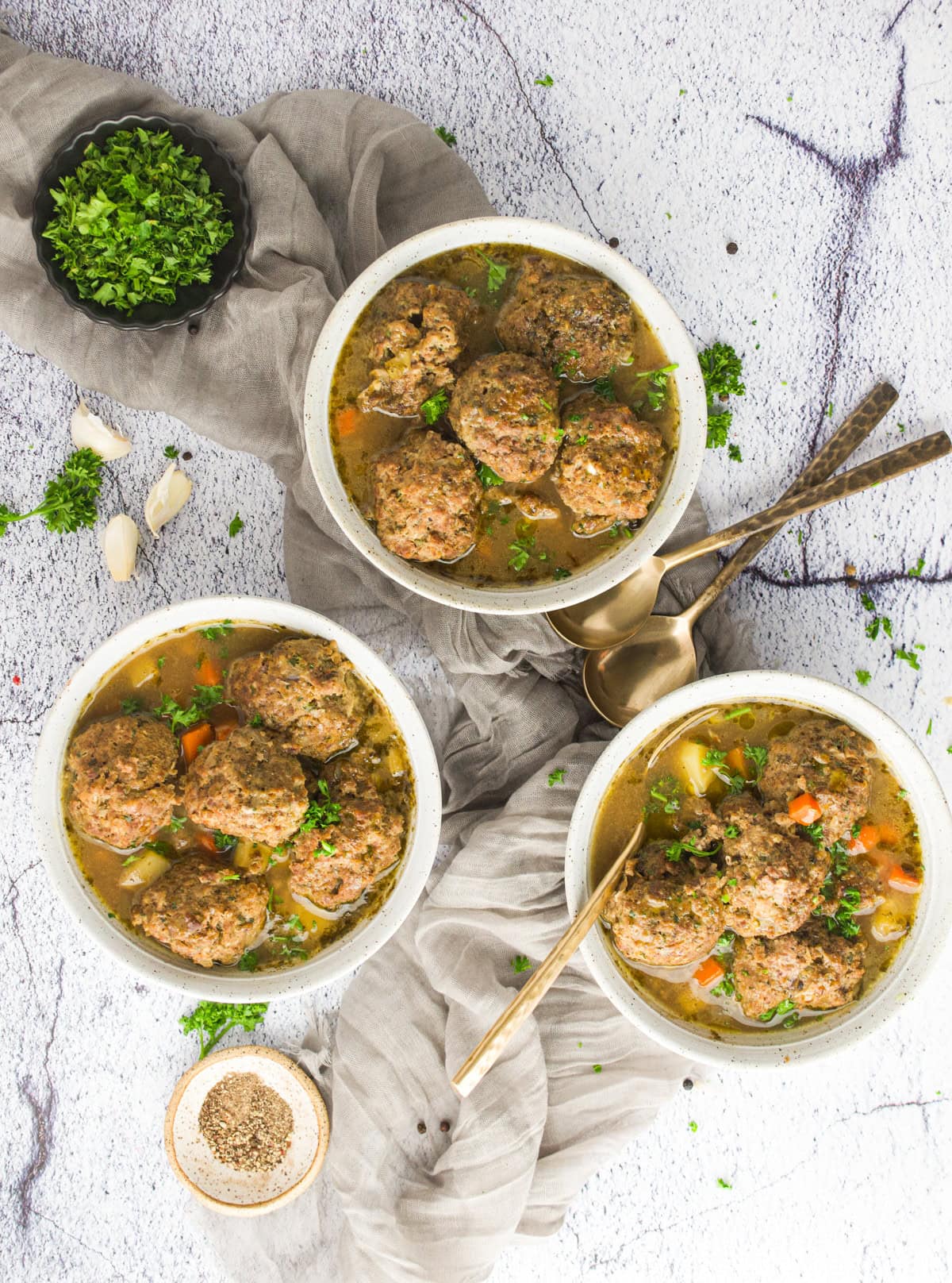Three bowls of Czech liver dumpling soup arranged on a light surface, surrounded by fresh parsley, garlic cloves, a bowl of pepper, and gold spoons on a gray linen cloth.