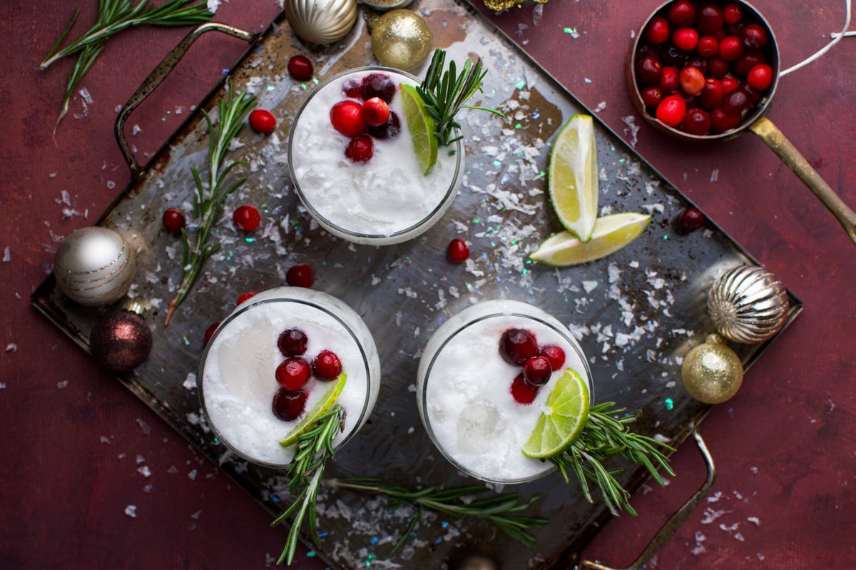 Three festive cocktails garnished with cranberries, lime slices, and rosemary sprigs on a tray, surrounded by Christmas ornaments, rosemary, cranberries, and lime wedges on a red surface with scattered faux snow.