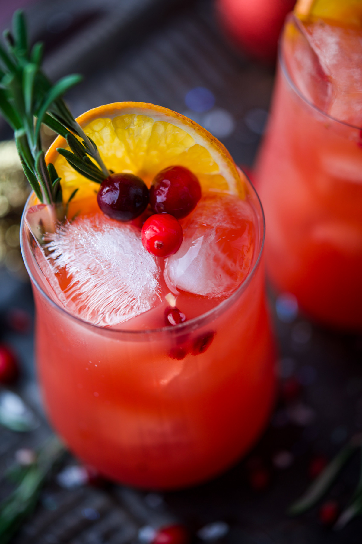 A glass of bright red cocktail with ice cubes, garnished with an orange slice, cranberries, and a sprig of rosemary. Another similar drink is blurred in the background.