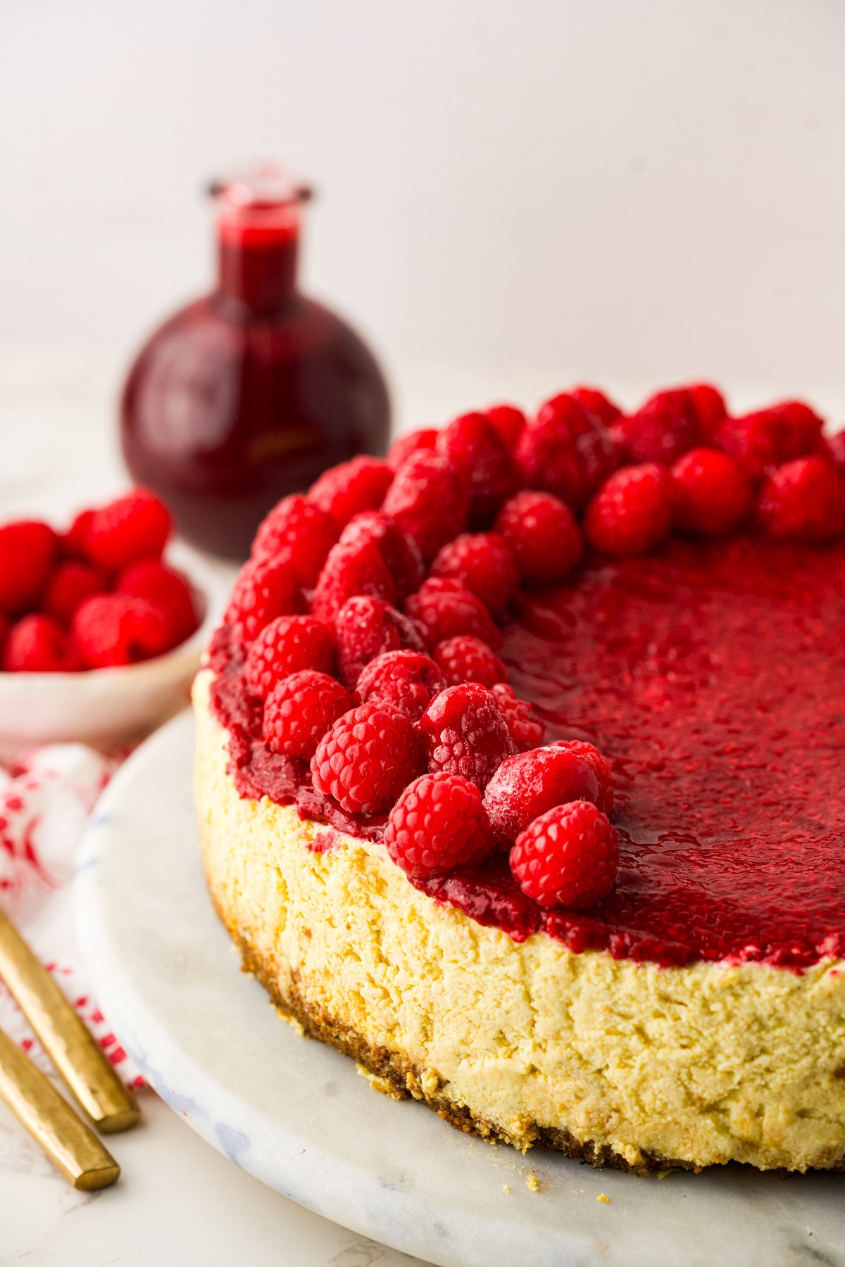 A cheesecake topped with a glossy raspberry sauce and fresh raspberries, set on a marble platter. A bowl of raspberries and a jar of raspberry sauce are in the background. Gold utensils are beside the cheesecake.