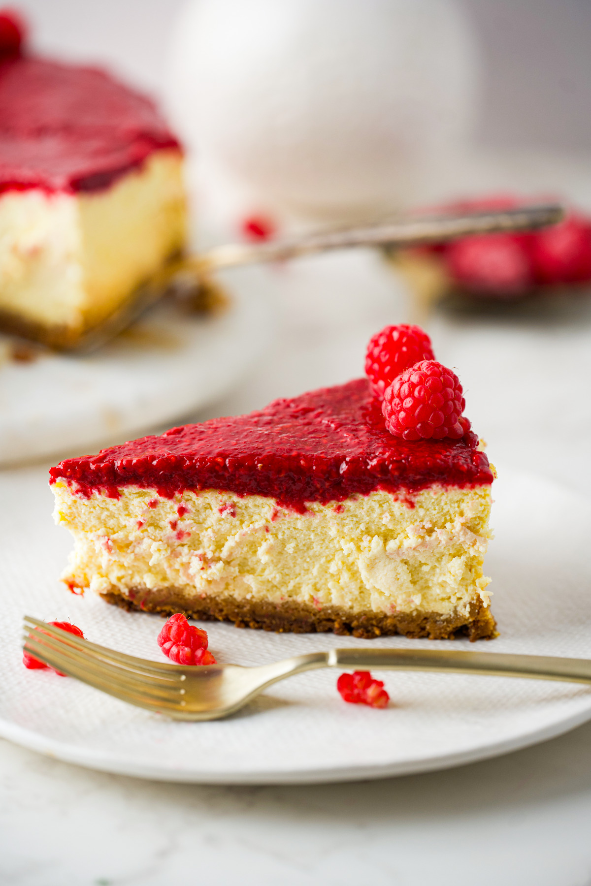 A slice of polish raspberry cheesecake topped with a layer of raspberry sauce and fresh raspberries sits on a white plate next to a fork, with another slice and some blurred raspberries in the background.
