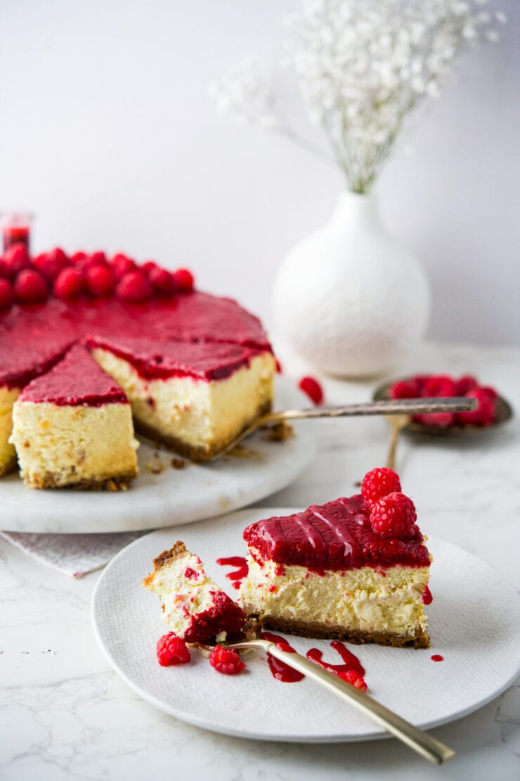 A slice of Polish raspberry cheesecake topped with raspberry sauce and fresh raspberries is served on a plate with a fork; the rest of the cheesecake and a vase of white flowers are in the background.