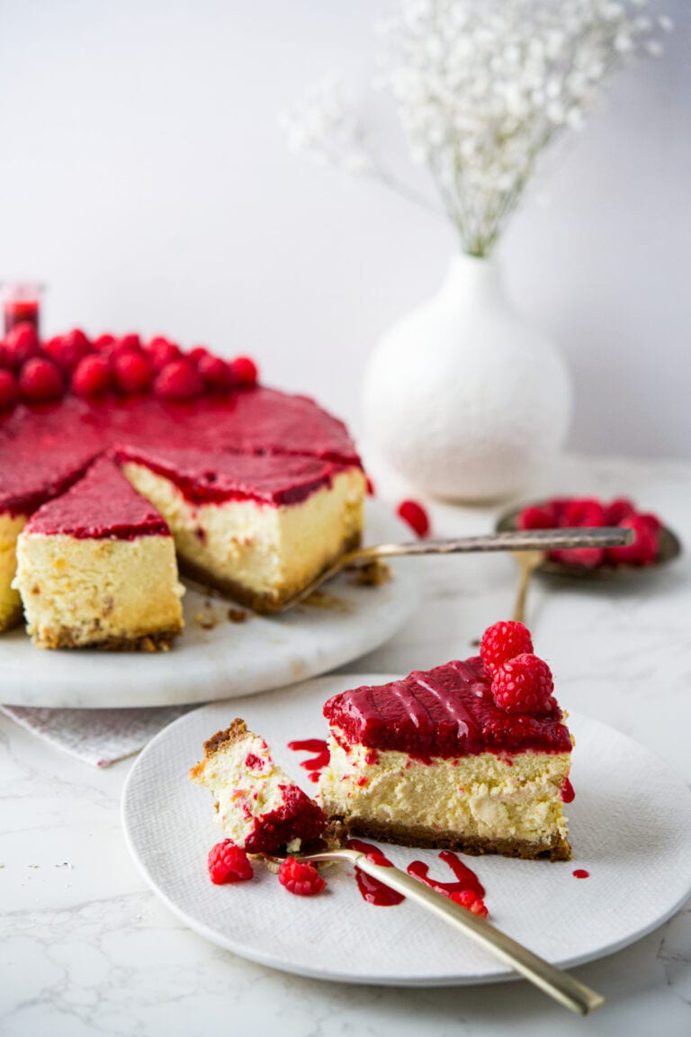 A slice of Polish raspberry cheesecake topped with raspberry sauce and fresh raspberries is served on a plate with a fork; the rest of the cheesecake and a vase of white flowers are in the background.
