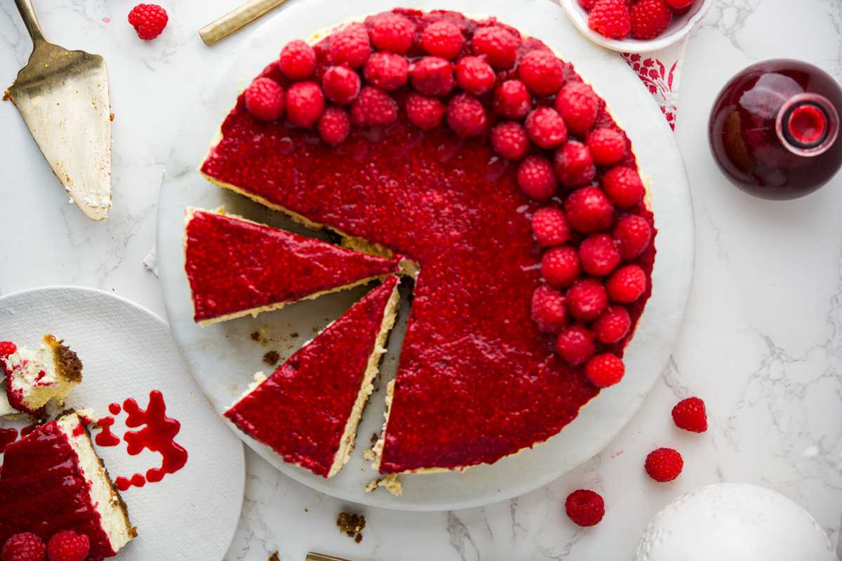 A polish raspberry-topped cheesecake with a bright red raspberry glaze, partially sliced, sits on a white plate. Fresh raspberries and a serving utensil are nearby on a marble surface.