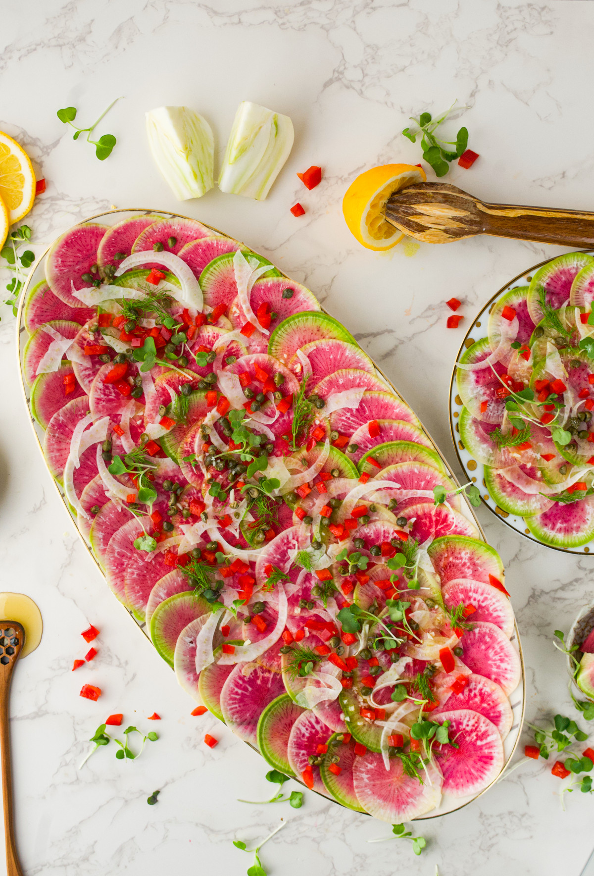 A large oval platter with Watermelon Radish Carpaccio topped with fennel, microgreens, diced red peppers, and herbs on a marble surface, with lemon wedges and utensils nearby.