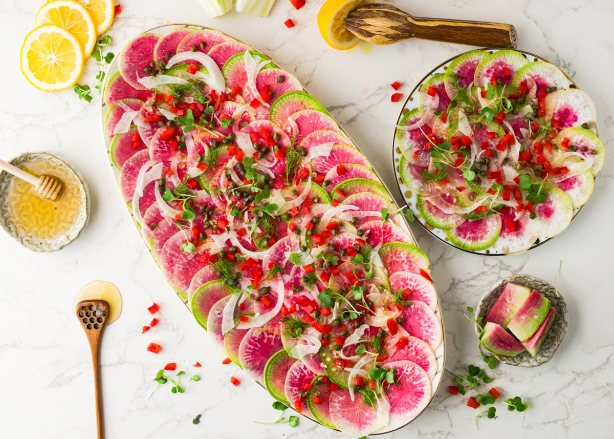 Oval and round plates display thinly sliced watermelon radishes arranged in a circular pattern, garnished with microgreens, diced red peppers, and onion slices. Lemon slices, honey, and serving utensils are nearby.