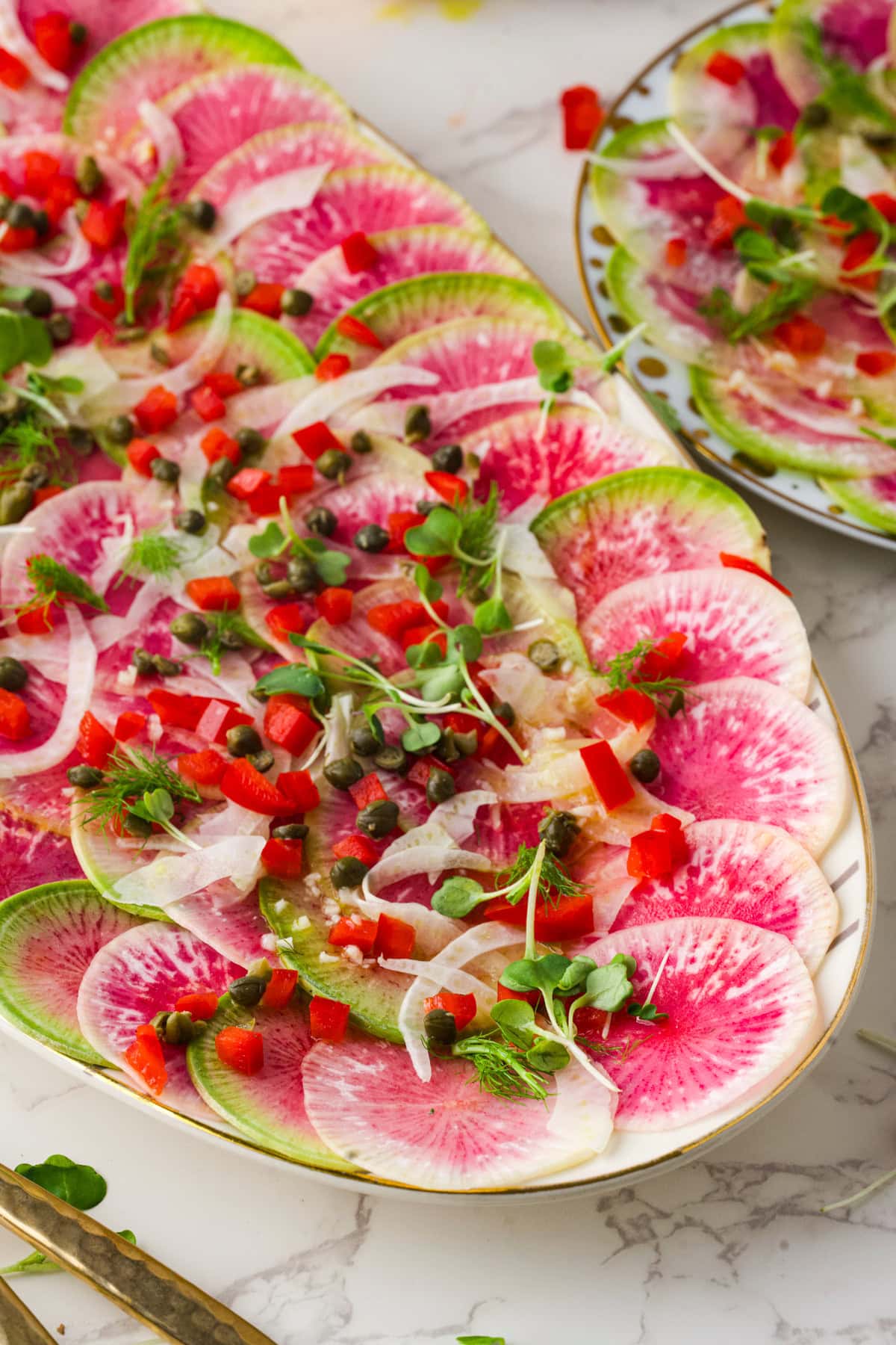 Thinly sliced watermelon radishes arranged on a platter, topped with fennel, capers, microgreens, and diced red peppers, creating a vibrant and colorful salad on a marble surface.