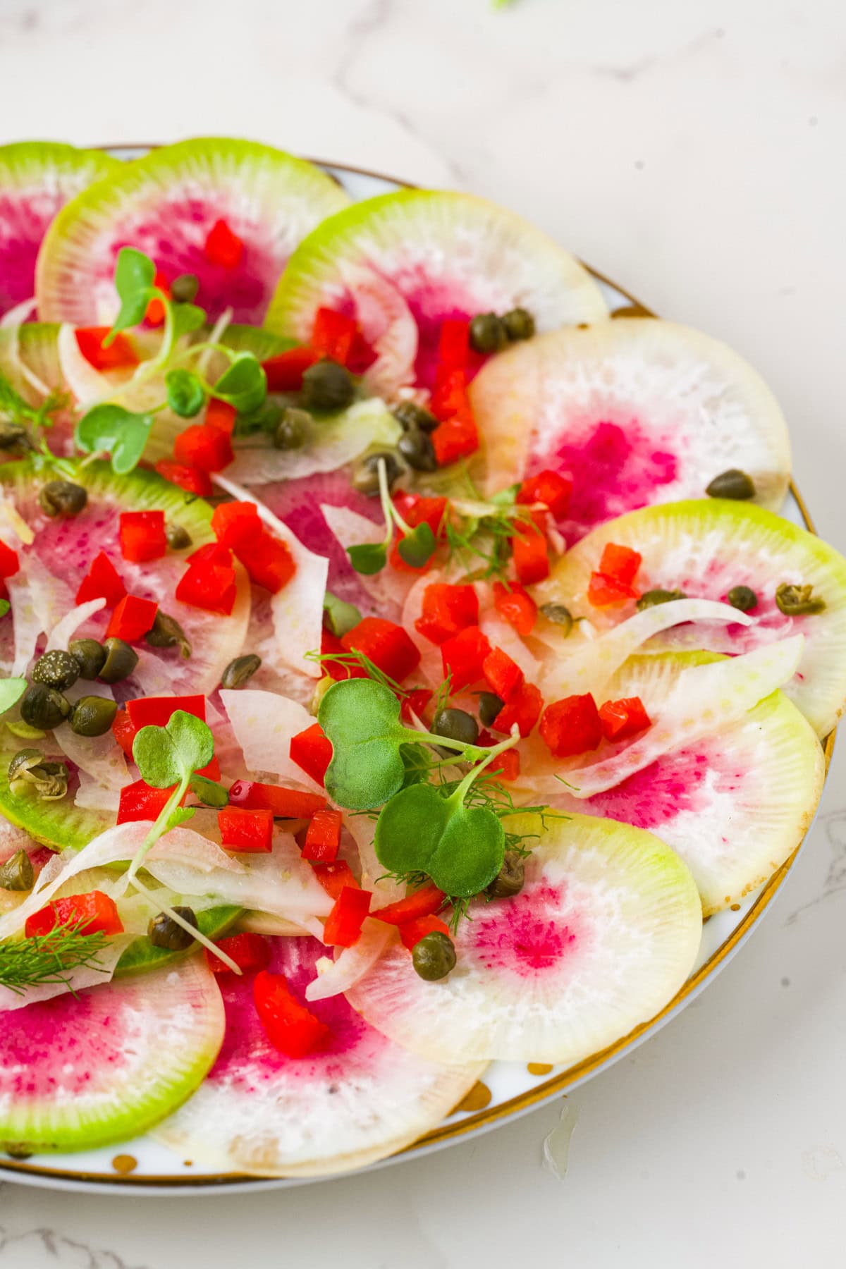 A plate of thinly sliced watermelon radishes topped with diced red bell peppers, microgreens, capers, and thinly sliced onions, arranged on a white surface.