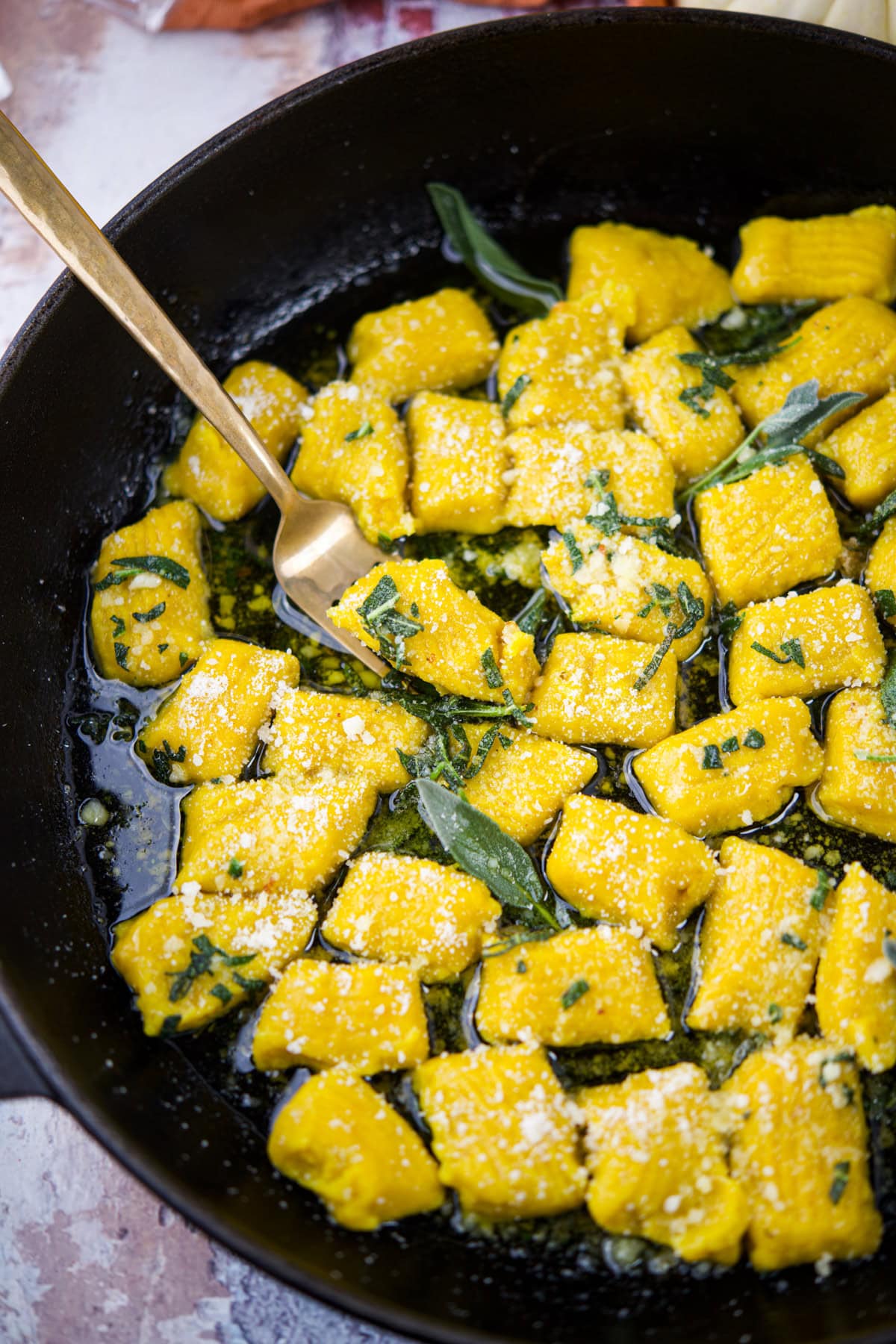 A skillet filled with golden-yellow gnocchi tossed in a butter and sage sauce, sprinkled with grated cheese. A gold fork rests in the pan among the gnocchi pieces.