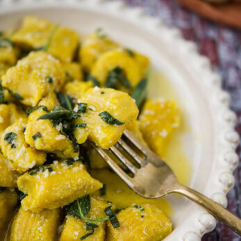 A plate of golden pumpkin gnocchi topped with fresh sage and a buttery sauce, served with a fork on a decorative white dish. The gnocchi are soft and coated in sauce, and a gourd is blurred in the background.